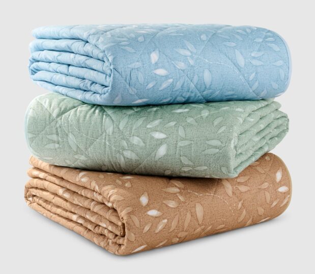 A stack of the Mary Jane Quilted bedspreads are shown here. It features all three colorways, Almond, Mineral Green and Slate Blue.